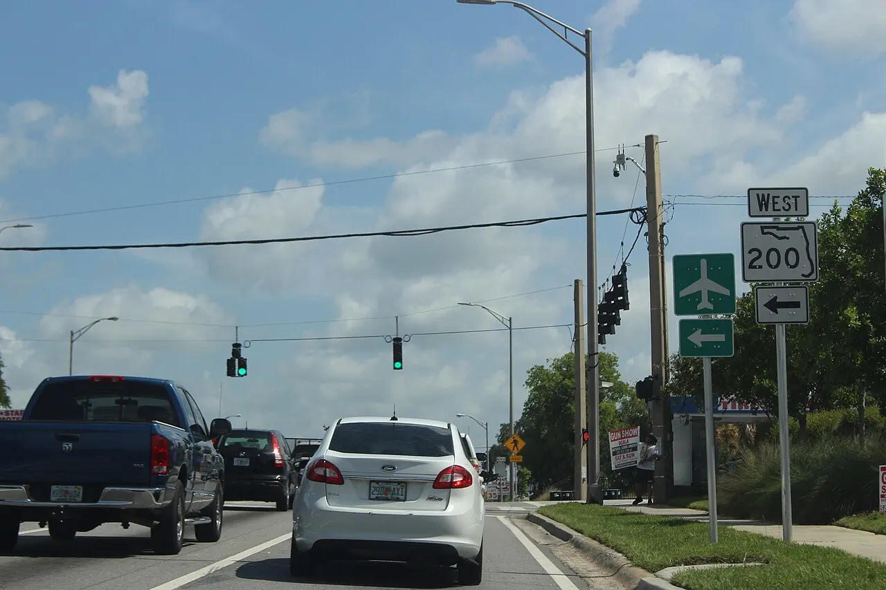 A wide multi-lane road on the south-west side of Ocala, with a green highway sign and pines on the verge.